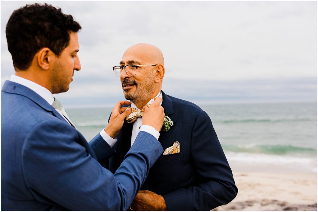 A groom straightens his dad's tie before family photos on the beach.