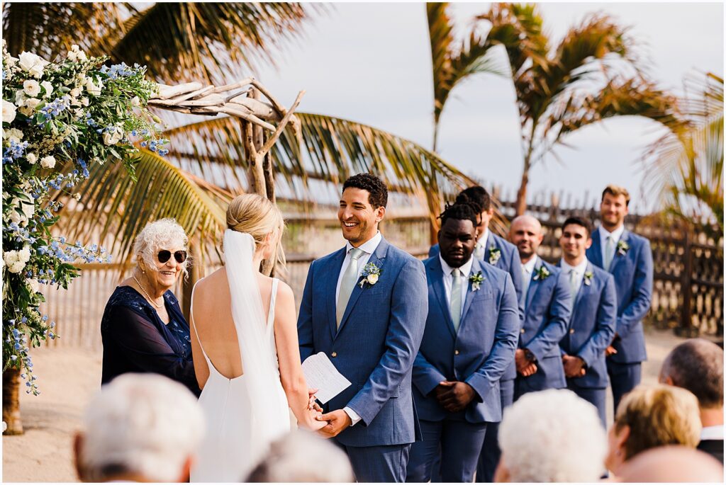 A groom smiles at a bride during a beach wedding ceremony.