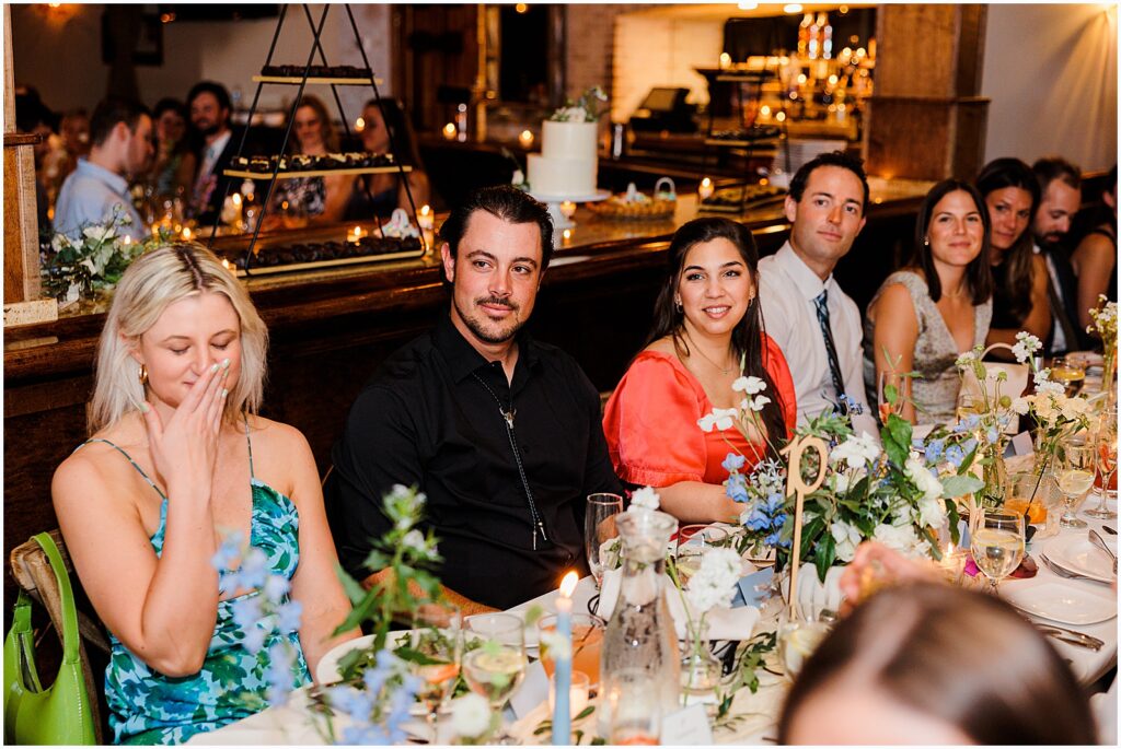 Wedding guests sit at a long table listening to speeches.