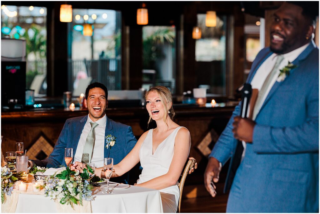 A bride and groom laugh during the best man speech at their wedding reception.