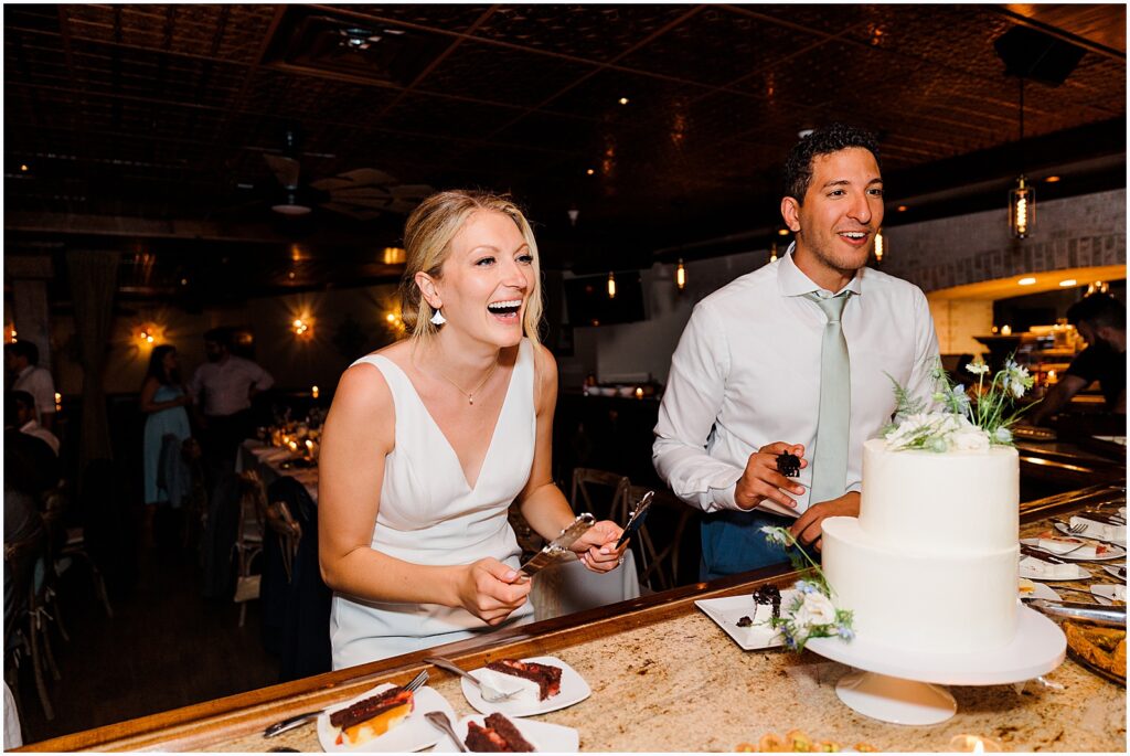 A bride and groom laugh beside a tiered wedding cake.