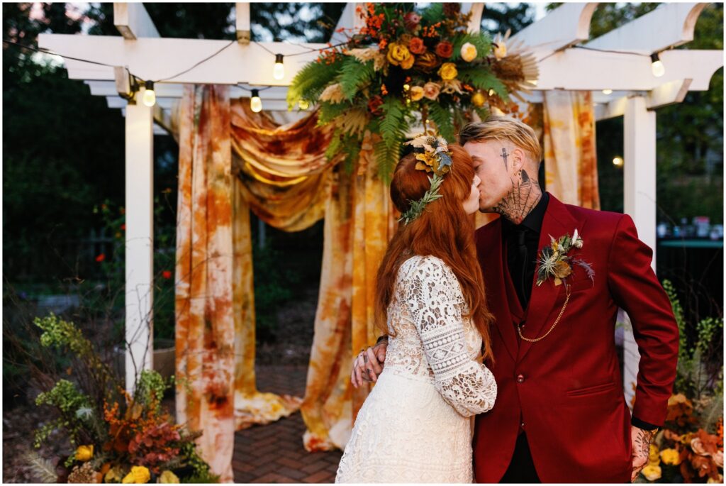 A bride and groom kiss under a floral wedding arch.