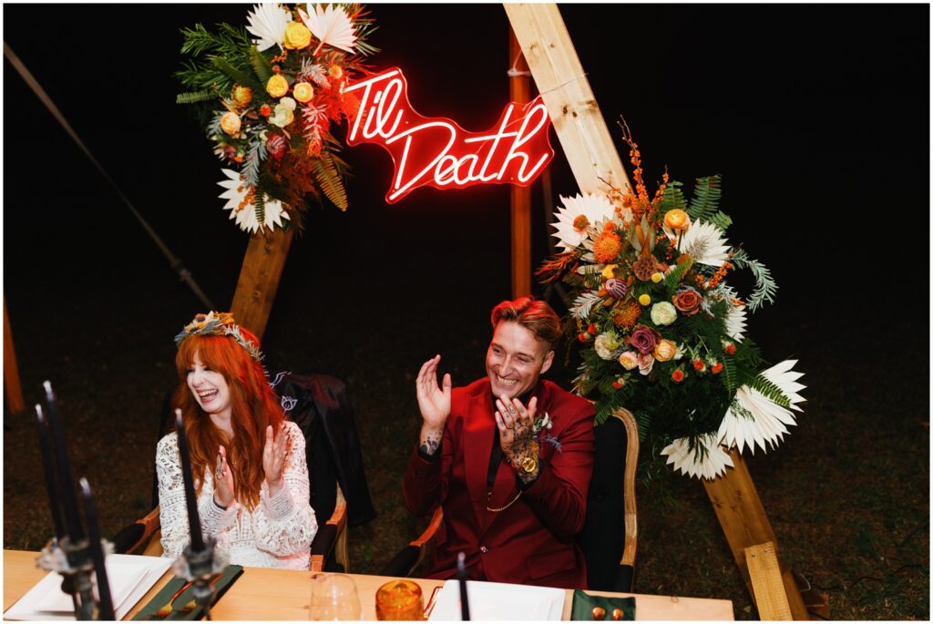 A bride and groom sit at a table under a neon sign that reads "til death."