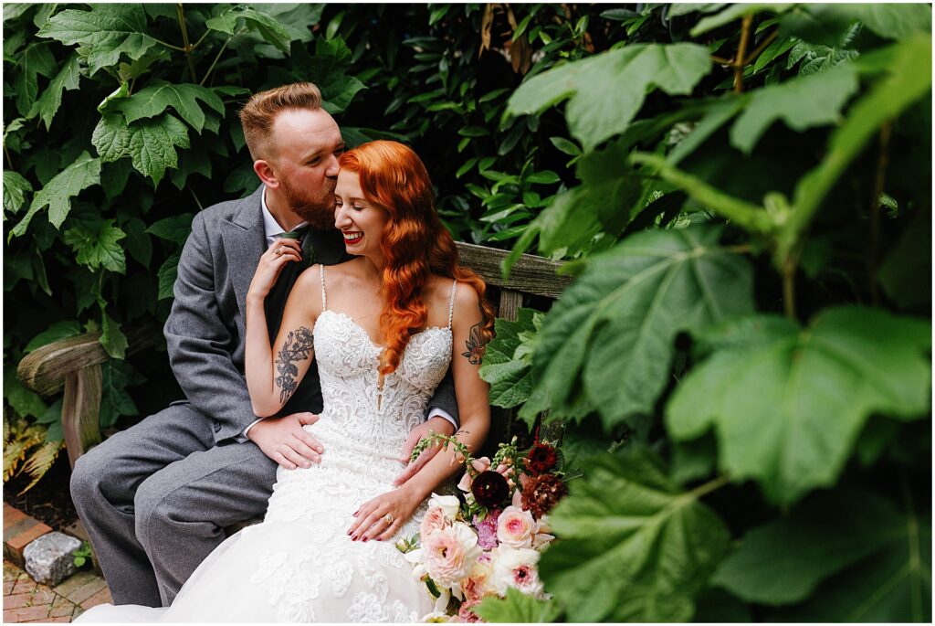 A groom kisses a bride's cheek on a bench in the herb garden at the Mutter Museum.