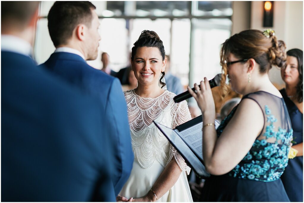 A bride smiles at a groom while an officiant reads a wedding script in Watermark Asbury Park.
