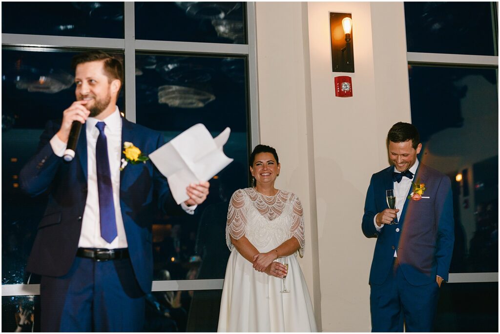 A bride and groom laugh as a best man gives a speech inside Watermark.