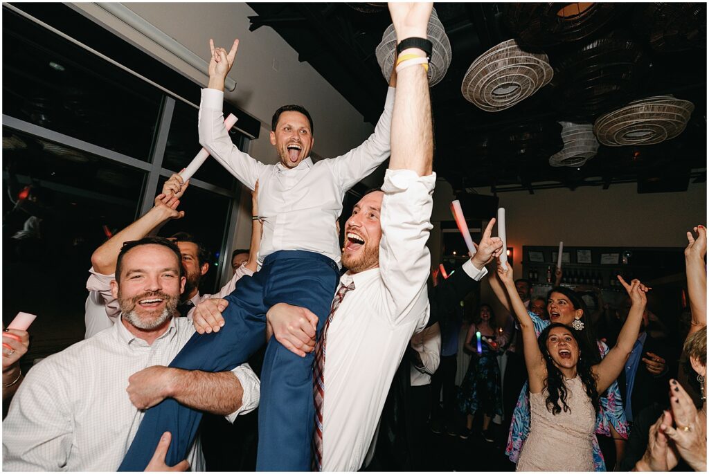 Wedding guests lift a groom on their shoulders.