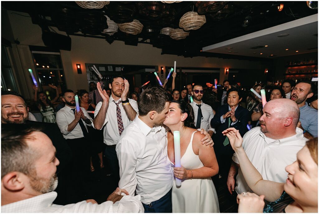 On a dance floor, a bride and groom kiss while wedding guests cheer.
